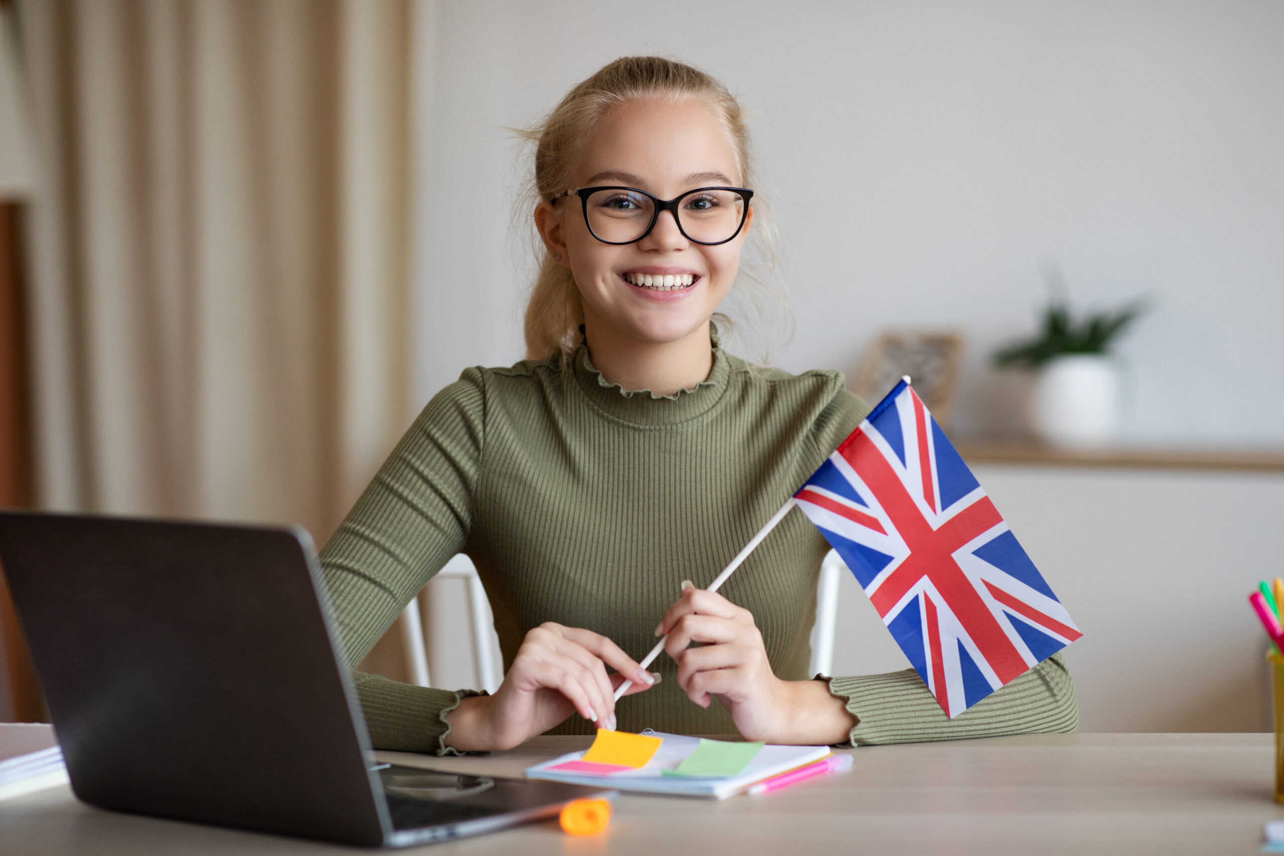 Une jeune fille assise à un bureau tient un drapeau anglais dans ses mains en souriant.