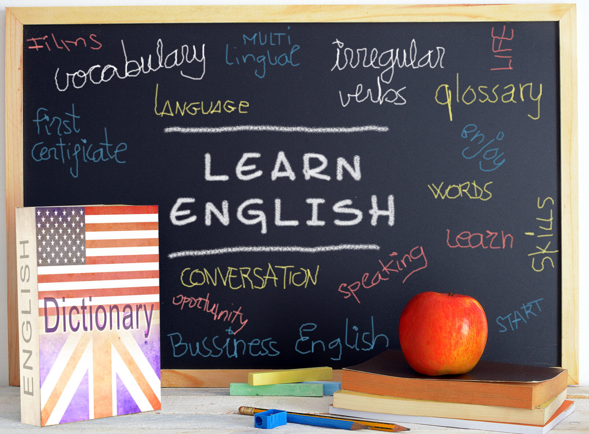 Un tableau noir avec des mots en anglais est posé sur une table avec un dictionnaire d’anglais et des fournitures scolaires.